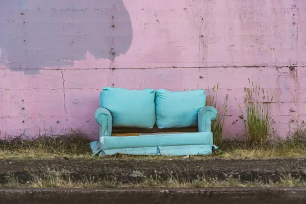 Worn turquoise loveseat left on a curb against a pink wall