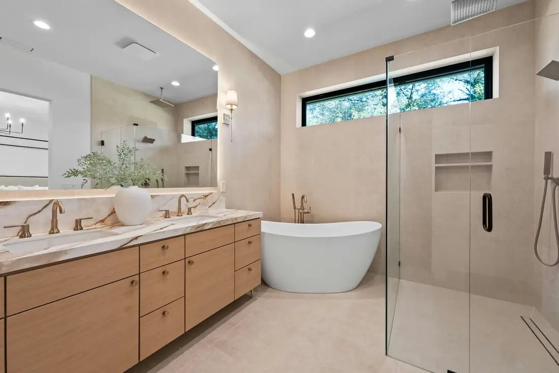 Warm primary bathroom with oak vanity, freestanding tub and glass shower